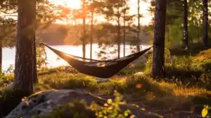 En hängmatta hänger mellan två träd i en solbelyst skog, omgiven av klippor och grönska. Solen går ner och kastar ett varmt gyllene sken över scenen, med en lugn vattenmassa i bakgrunden.