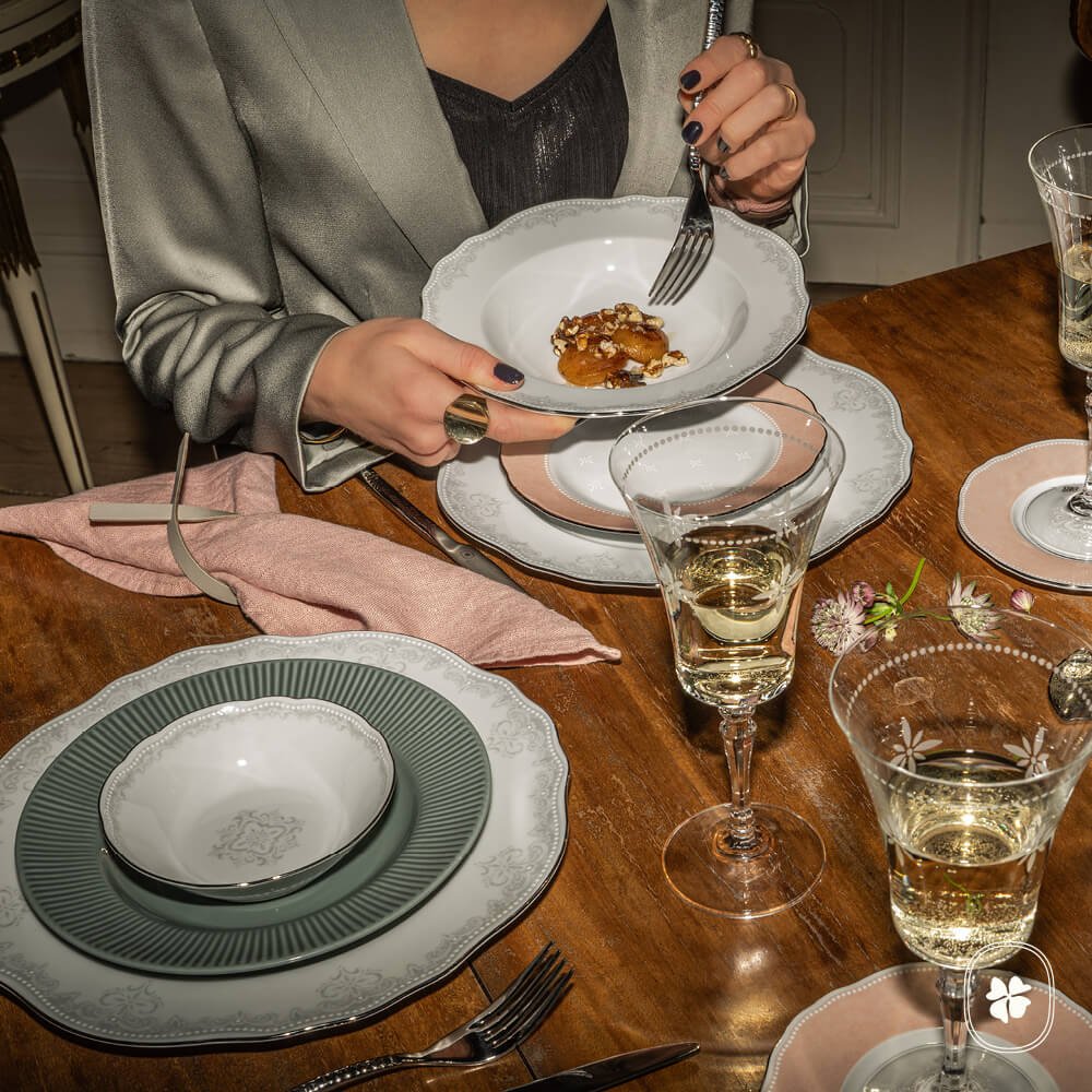 En kvinna i satinblåsa sitter vid ett träbord dukat med eleganta tallrikar, kristallglas med vitt vin och rosa servetter. Hon håller i en skål med mat och en gaffel, redo att äta. Blomsterdekorationer är arrangerade på bordet.