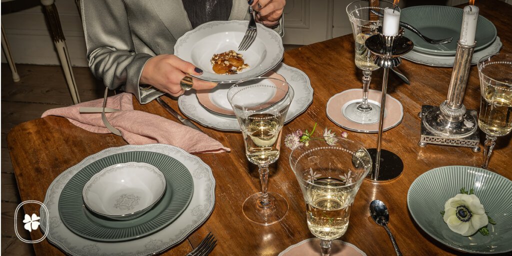 En person i grå klädsel äter dessert ur en vit skål vid ett elegant bord med levande ljus, dukat med vinglas, tallrikar, rosa servetter och silverbestick på en träyta.