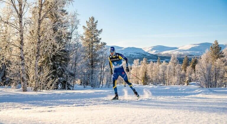 En längdskidåkare i blågul dräkt glider fram genom ett snöigt, solbelyst landskap med frostnupna träd och avlägsna berg under en klarblå himmel.